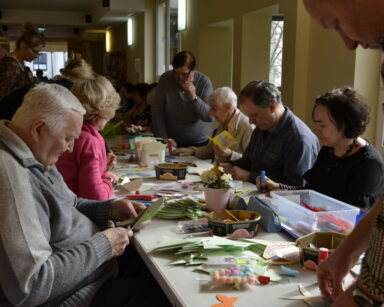 Sala. Stół. Grupa osób pracuje nad wykonaniem wielkanocnych kartek. Na stole przybory do wykonania kartek.