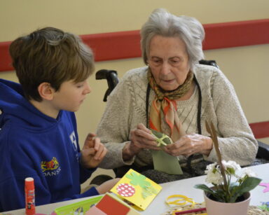 Zbliżenie. Seniorka trzyma w ręku zieloną kartkę z kokardką. Obok siedzi chłopiec i pomaga wyklejać kartki.