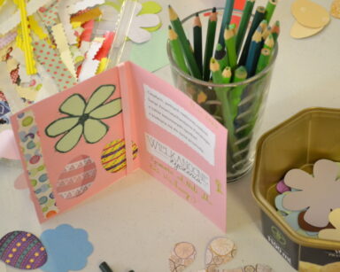 Zbliżenie. Stół. Na stole różowa kartka z życzeniami, kwiatkiem i pisankami. Obok ozdoby do wyklejania kartek.