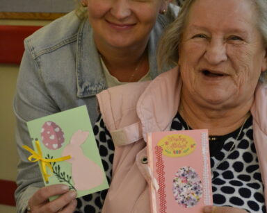 Zbliżenie. Dwie kobiety pozują do zdjęcia. Seniorka trzyma dwie kartki wielkanocne w dłoniach. Kobiety uśmiechają się.