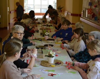 Sala. Długi stół, przy nim dzieci i seniorzy przygotowują wielkanocne kartki. Na stole nożyczki, kartki, klej, ozdoby.