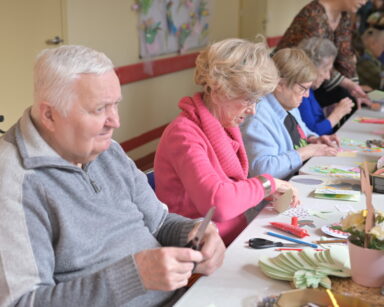 Sala. Grupa seniorów przy stole wykleja kartki. Na stole akcesoria do wykonania świątecznych kartek.