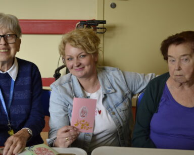 Zbliżenie. Trzy kobiety pozują do zdjęcia. Jedna z kobiet trzyma różową wyklejana kartkę wielkanocną.