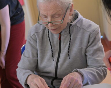 Zbliżenie. Seniorka w szarej bluzie siedzi przy stole. Wykleja kolorowymi motylkami, zajączkami świąteczną kartkę.