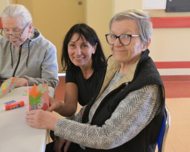 Zbliżenie. Młoda kobieta i dwie seniorki pozują przy stole do zdjęcia. Seniorka trzyma w ręku kolorową wyklejana kartkę.