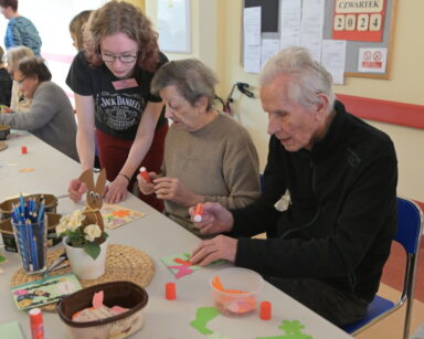 Zbliżenie. stół. Wolontariuszka pomaga parze seniorów przy wyklejaniu i wypisaniu kartki świątecznej.