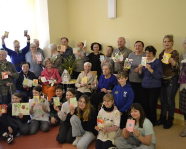 Sala. Grupa seniorów, opiekunów i dzieci pozuje do wspólnego zdjęcia. Wszyscy trzymają kolorowe kartki wielkanocne.