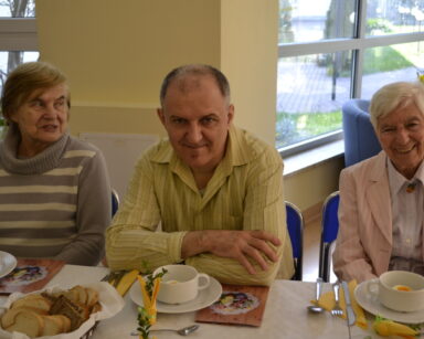 Zbliżenie. Troje seniorów uśmiecha się do zdjęcia. Seniorzy siedzą przy stole. Na stole ozdoby, talerze, babki, pieczywo.