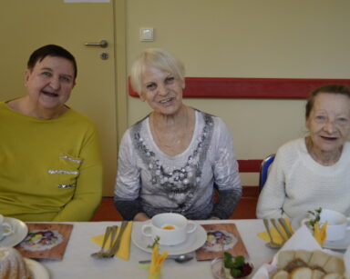 Zbliżenie. Trzy uśmiechnięte seniorki siedzą przy wielkanocnym stole i pozują do zdjęcia. Na stole wielkanocne jedzenie.