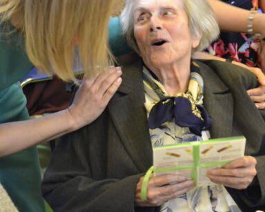 Zbliżenie. Kobieta pochyla się do uśmiechniętej seniorki, siedzącej na wózku. Seniorka w dłoniach trzyma czekoladki.
