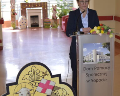 Zbliżenie. Pani Prezydentka Sopotu stoi przy mównicy. Na niej napis Sopockie Centrum Seniora. Obok figurka Baranka Bożego.