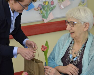 Zbliżenie. Kobieta pochyla się nad seniorką i wręcza jej torbę z napisem Sopockie Centrum Seniora i tulipana.