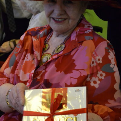 Zbliżenie. Seniorka w czerwonym kapeluszu i kwiecistej sukience pozuje do zdjęcia. W dłoni trzyma pudełko czekoladek.