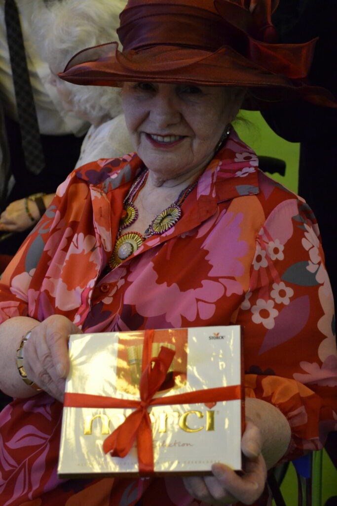 Zbliżenie. Seniorka w czerwonym kapeluszu i kwiecistej sukience pozuje do zdjęcia. W dłoni trzyma pudełko czekoladek.