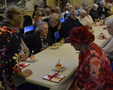 Sala. Dwa długie stoły, przy nich siedzą seniorzy. Na stołach ciasto. czekoladki. W tle grupa ludzi.