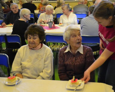 Zbliżenie. Uśmiechnięta wolontariuszka podaje ciasto seniorce. Obok siedzi seniorka i patrzy na ciasto. W tle grupa seniorów.