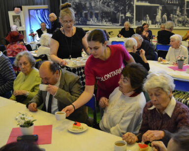 Sala. Grupa seniorów siedzi przy stołach. Wolontariuszki i kobieta z tacą rozdają napoje w filiżankach.