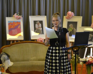Scena. Kobieta stoi przy mikrofonie, w ręku trzyma kartkę. W tle kanapa, kwiaty, obraz i zdjęcia seniorek.