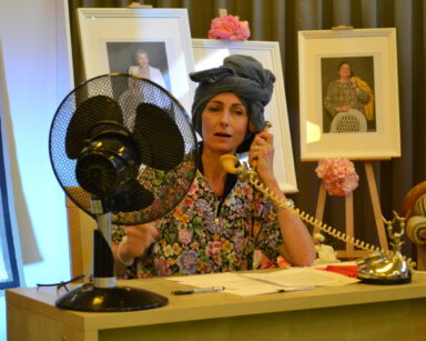 Scena. Kobieta w szlafroku i ręczniku na głowie siedzi przy biurku, w ręku trzyma telefon przy uchu. W tle kanapa, obraz, zdjęcia.