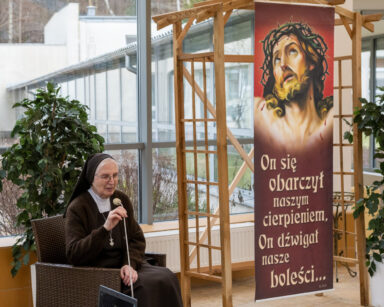 Sala. Siostra zakonna siedzi na krześle w ręku ma mikrofon. Obok zdjęcie Chrystusa w koronie cierniowej.