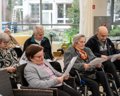 Sala. Grupa osób siedzi na fotelach. Seniorzy trzymają kartki. W tle okna i widok na ogród.