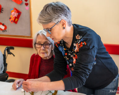 Zbliżenie. Kobieta w koszuli w kwiaty pomaga przy stole seniorce pleść wianek i ozdabiać małymi kolorowymi jajeczkami.