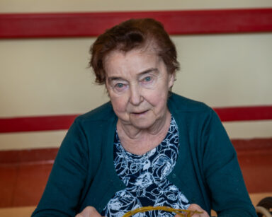 Zbliżenie. Uśmiechnięta seniorka pozuje do zdjęcia. Siedzi przy stole, w ręku trzyma uplecione wianki z gałązek.