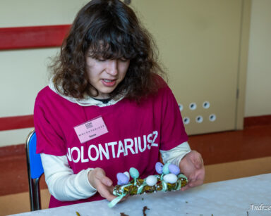 Zbliżenie. Wolontariuszka siedzi przy stole. W rękach trzyma wianek z wierzby ozdobiony kolorowymi małymi jajeczkami.