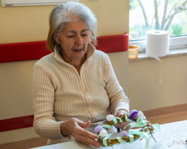 Zbliżenie. Seniorka siedzi przy stole. W rękach trzyma wianek ozdobiony kolorowymi jajeczkami i zieloną wstążką.