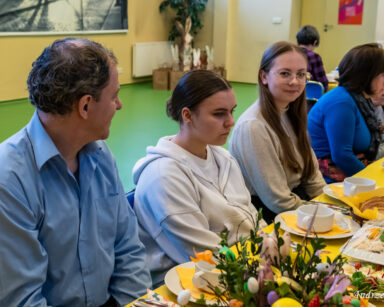 Sala. Grupa osób siedzi przy stole wielkanocnym. Na stole chleb, talerze, jajeczka, sałatka stroik wielkanocny.