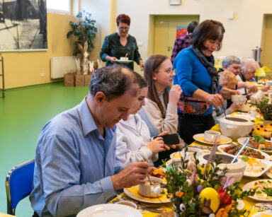 Sala. Grupa osób siedzi przy wielkanocnym stole. Seniorka z wazy wlewa na talerz żur. W tle kobieta niesie w ręku filiżankę.