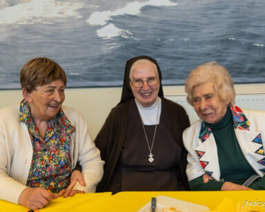 Zbliżenie. Trzy seniorki pozują do zdjęcia. Kobiety uśmiechają się, siedzą przy stole. W tle czarno białe zdjęcie.