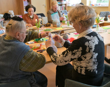 Sala. Grupa seniorów przy stole robi wielkanocne zajączki ze skarpet, ryżu, kokardek i kolorowych koralików.