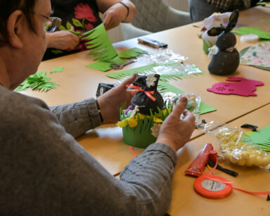 Zbliżenie. Na stole kolorowe ze skarpet zrobione zajączki, zielona trawa z papieru, klej, nożyczki, ozdobne jajeczka.