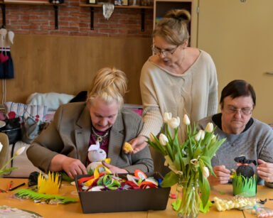 Zbliżenie. Dwie seniorki ozdabiają zające, obok kobieta podaje seniorce żółty kwiatek. Na stole wazony z tulipanami.