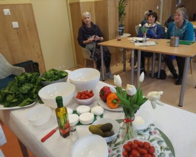 Sala. Na stole miski, warzywa, oliwa, sery, pomidorki. W tle przy stolikach siedzą seniorzy.