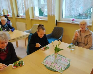 Zbliżenie. Trzy seniorki siedzą przy stole i jedzą warzywną sałatkę. Na stole serweta, wazon z kwiatami.