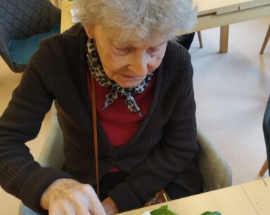 Zbliżenie. Seniorka siedzi przy stoliku. Trzyma widelec w sałatce. W tle seniorzy przy stołach jedzą sałatki.