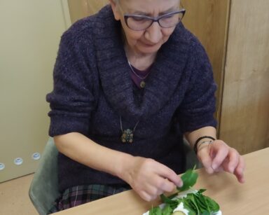 Zbliżenie. Seniorka siedzi przy stoliku i talerzyku z sałatką. Trzyma w palcach liście szpinaku.