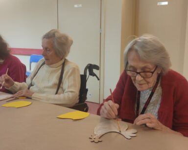 Korytarz. Pięć osób przy stole. Na stole szablony kurek, żółte kartki. Seniorki w dłoniach trzymają pędzle.