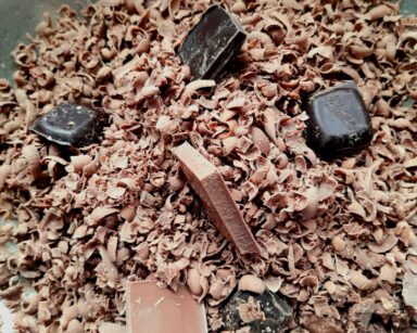 Zbliżenie. W metalowej, srebrnej misce leży tarkowana na drobno czekolada, obok leży kilka całych kostek czekolady.