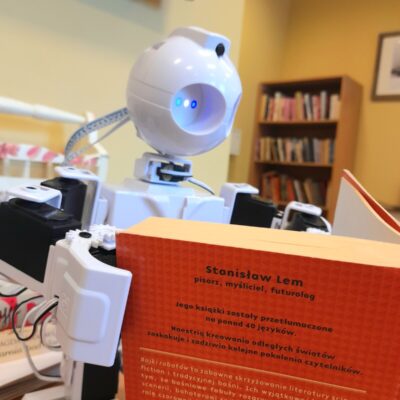 Zbliżenie. Robot JD trzyma w rączkach książkę. Stanisław Lem "Bajki Robotów". W tle regał z książkami.