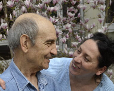 Ogród. Senior i kobieta siedzą na ławeczce. Kobieta uśmiecha się do mężczyzny. W tle drzewo magnolii.