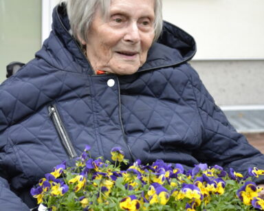 Zbliżenie. Uśmiechnięta seniorka trzyma na kolanach kartonik z kwiatami i napisem 