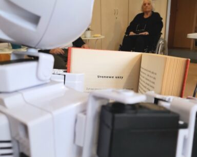 Zbliżenie. Na stoliku robot trzyma otwartą książkę. W tle seniorka w ciemnym ubraniu siedzi na wózku i patrzy na robota.