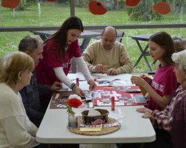 Sala. Dwie młode wolontariuszki pomagają zrobić patriotyczny kolaż. Przy stole siedzi pięciu seniorów.
