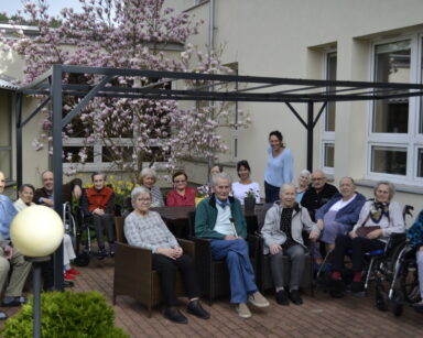 Ogród. Zbliżenie. Grupa osób pozuje do zdjęcia. W tle drzewo magnolii o różowych kwiatach.