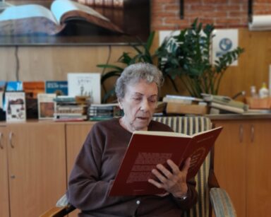 Zbliżenie. Seniorka siedzi na fotelu i trzyma w ręku brązową książkę. W tle na półce książki i obraz biblioteczki.