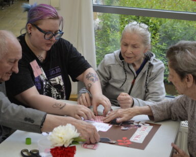 Zbliżenie. Wolontariuszka pomaga trójce seniorom wykleić patriotyczny kolaż. Na stole nożyczki, klej i wazon z kwiatami.