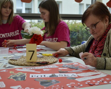 Zbliżenie. Seniorka wykleja kolaż. W tle nad kolażem pracują dwie wolontariuszki. Na stole numer 7, wycinanki, nożyczki.